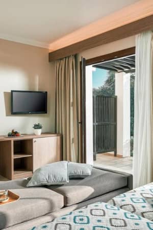 Luxurious hotel room at Royal Mare with a grey chaise lounge, flat-screen TV, round mirror, and access to private terrace through sliding doors.