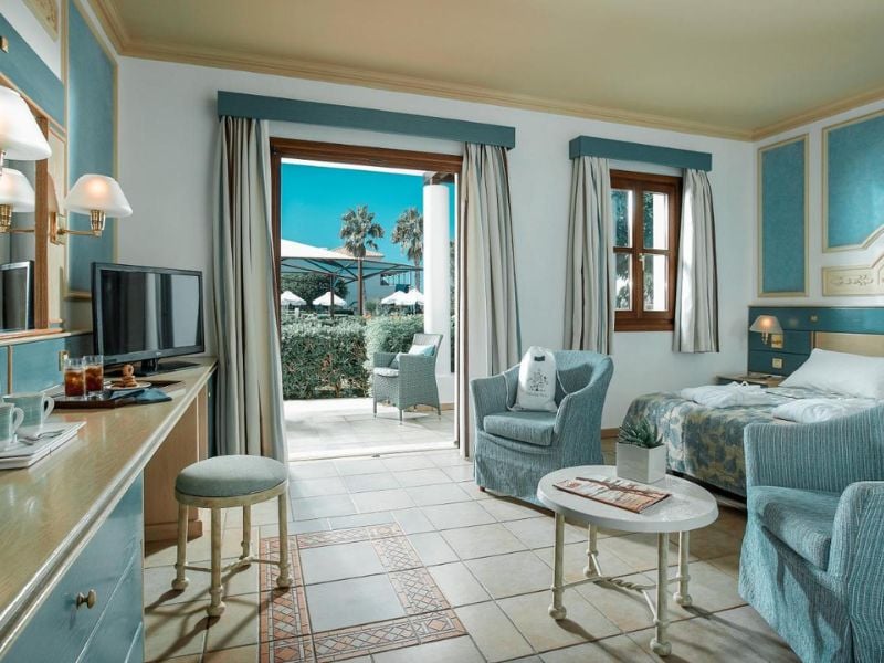 Luxurious hotel room with blue and white decor at Royal Mare, featuring a twin bed, armchair, TV, and garden view with palm trees and umbrellas.