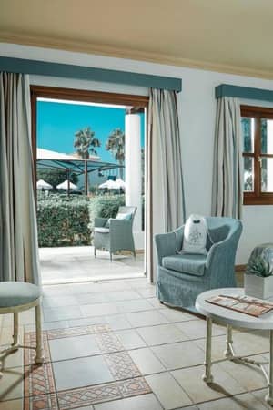 Luxurious hotel room with blue and white decor at Royal Mare, featuring a twin bed, armchair, TV, and garden view with palm trees and umbrellas.
