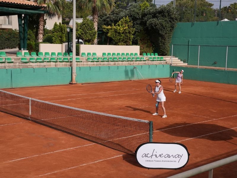 Two women playing tennis on a clay court at Royal Mare, with one serving and the other at the baseline. Surroundings include green fences and seating.