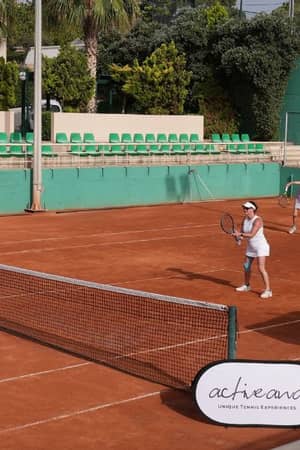 Two women playing tennis on a clay court at Royal Mare, with one serving and the other at the baseline. Surroundings include green fences and seating.