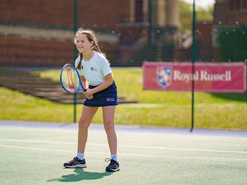 Royal Russell School student in ready position on outdoor tennis court with racket, wearing sports gear with school branding in the background.