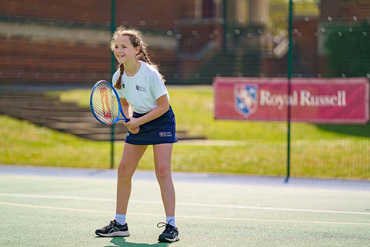 Royal Russell School student in ready position on outdoor tennis court with racket, wearing sports gear with school branding in the background.