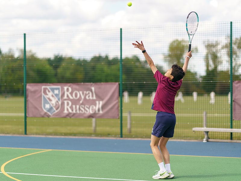Student serving tennis on the Royal Russell School tennis court with school branding and a scenic backdrop.
