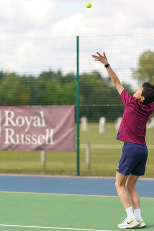 Student serving tennis on the Royal Russell School tennis court with school branding and a scenic backdrop.