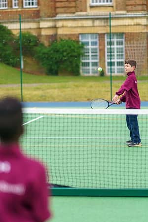 Two students playing tennis on an outdoor court at Royal Russell School, with a brick building in the background.