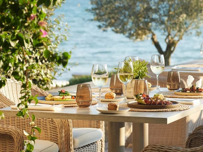 Outdoor dining table at Sani Beach with gourmet dishes, glasses of white wine, wicker chairs, and ocean view in the background.