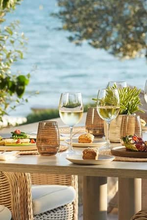 Outdoor dining table at Sani Beach with gourmet dishes, glasses of white wine, wicker chairs, and ocean view in the background.