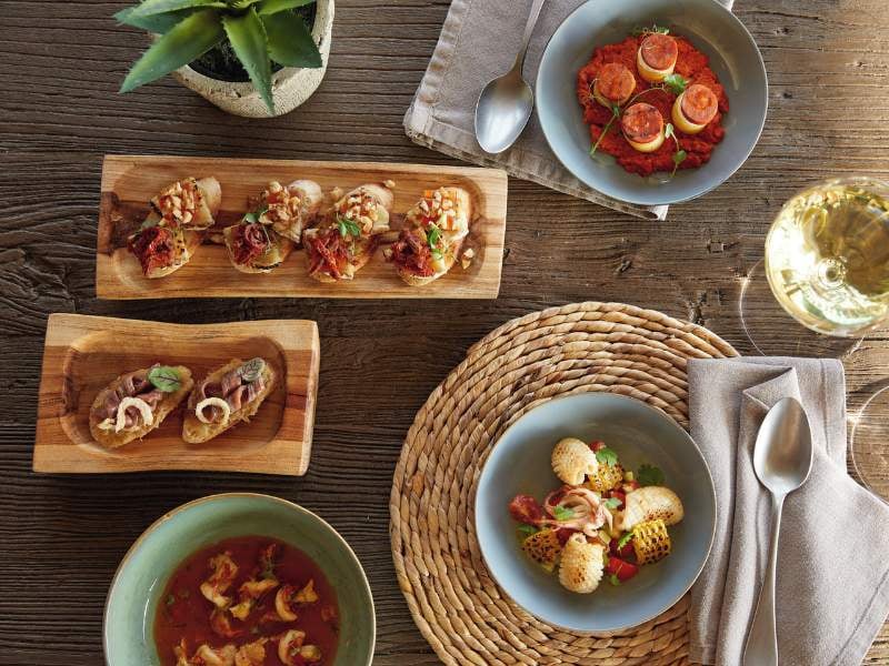 Mediterranean meal at Sani Beach with bruschetta, tomato-based dish, soup, and grilled shellfish on a rustic wooden table setting with a glass of white wine.