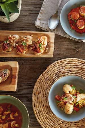 Mediterranean meal at Sani Beach with bruschetta, tomato-based dish, soup, and grilled shellfish on a rustic wooden table setting with a glass of white wine.