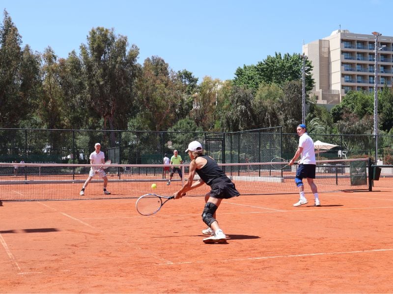 Tennis players on a red clay court at Sani Beach Resort with trees and buildings in the background.