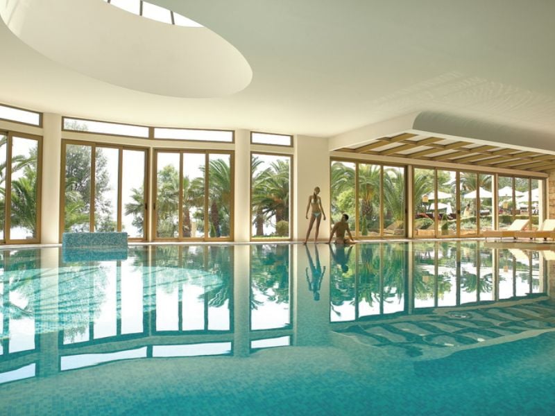 Luxurious indoor pool at Sani Beach with large windows showcasing views of palm trees and reflective blue water.