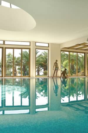 Luxurious indoor pool at Sani Beach with large windows showcasing views of palm trees and reflective blue water.