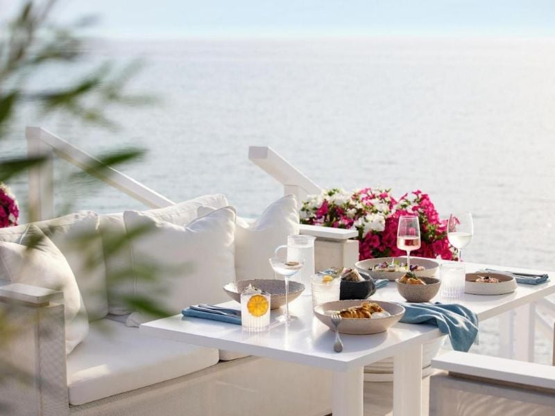 Outdoor dining area at Sani Beach with a sea view, white couch, and a table beautifully set with glassware and dishes, surrounded by vibrant pink flowers.