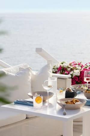 Outdoor dining area at Sani Beach with a sea view, white couch, and a table beautifully set with glassware and dishes, surrounded by vibrant pink flowers.