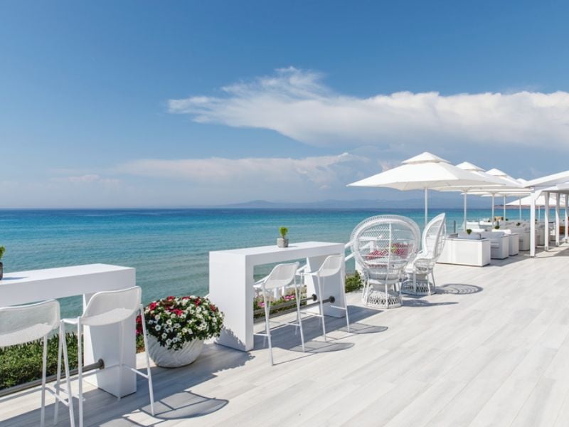 Outdoor seating at Sani Beach Resort, with white tables and umbrellas overlooking the turquoise sea.