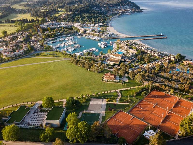 Aerial view of Sani Resort, Halkidiki, featuring a marina, sandy beach, tennis courts, and lush landscapes by the Aegean Sea.