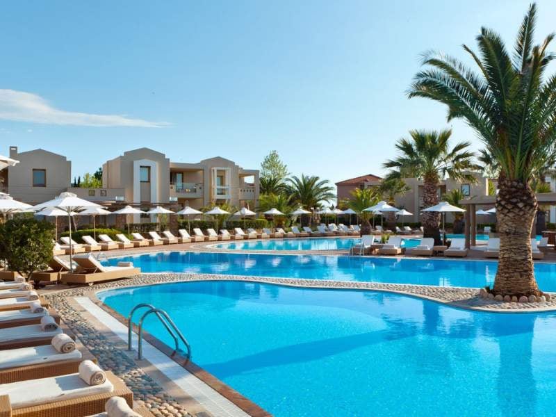 Luxurious outdoor pool at Sani Beach with sun loungers, palm trees, and modern buildings in the background.