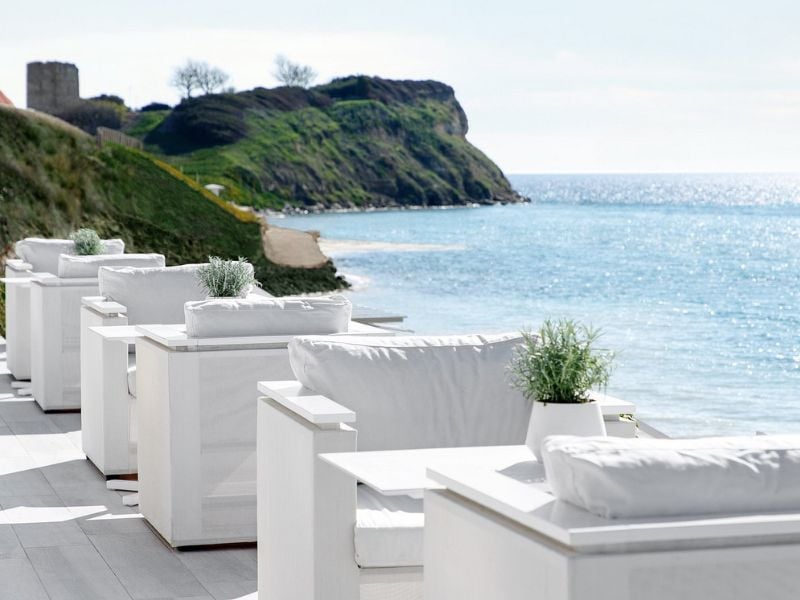 Outdoor dining area at Sani Beach with white seating and tables, overlooking the sea and a grassy hill under a clear sky.