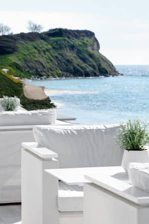 Outdoor dining area at Sani Beach with white seating and tables, overlooking the sea and a grassy hill under a clear sky.