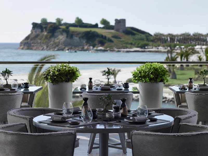 Outdoor terrace of Sani Beach Resort restaurant with elegantly set tables, overlooking a sandy beach, blue water, and an island with greenery and a historic tower.