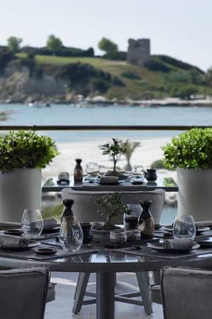 Outdoor terrace of Sani Beach Resort restaurant with elegantly set tables, overlooking a sandy beach, blue water, and an island with greenery and a historic tower.