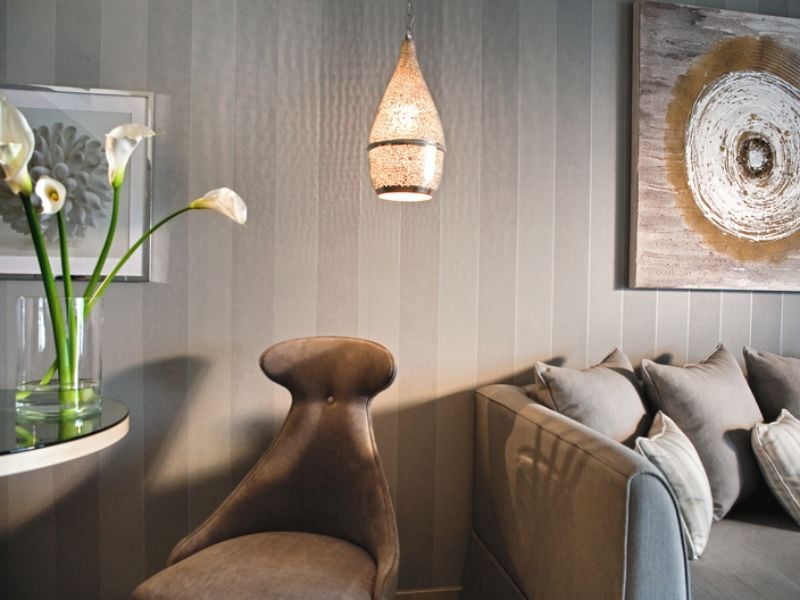 Interior view of a Sani Beach room with grey striped wall, plush grey chair, table with calla lilies, sofa with cushions, and decorative art with hanging light.