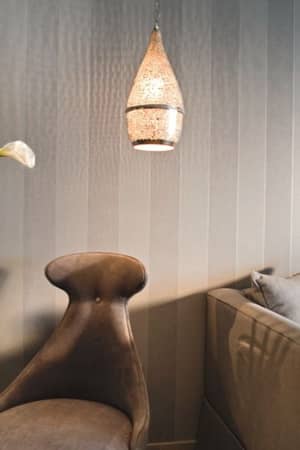 Interior view of a Sani Beach room with grey striped wall, plush grey chair, table with calla lilies, sofa with cushions, and decorative art with hanging light.