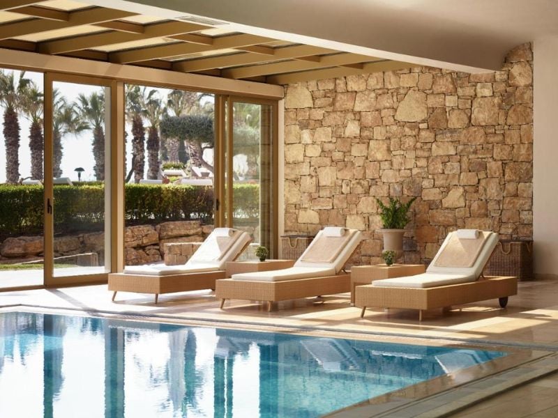 Spa lounge at Sani Beach with sun loungers by an indoor pool, surrounded by large glass doors showing garden and palms.
