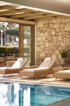 Spa lounge at Sani Beach with sun loungers by an indoor pool, surrounded by large glass doors showing garden and palms.