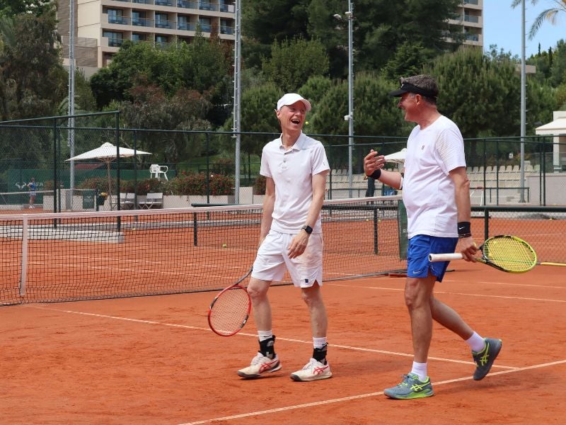 Two men in athletic gear playing tennis on a clay court at Sani Beach resort, surrounded by greenery and resort buildings.