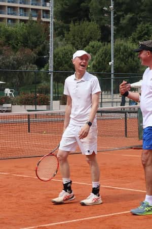 Two men in athletic gear playing tennis on a clay court at Sani Beach resort, surrounded by greenery and resort buildings.