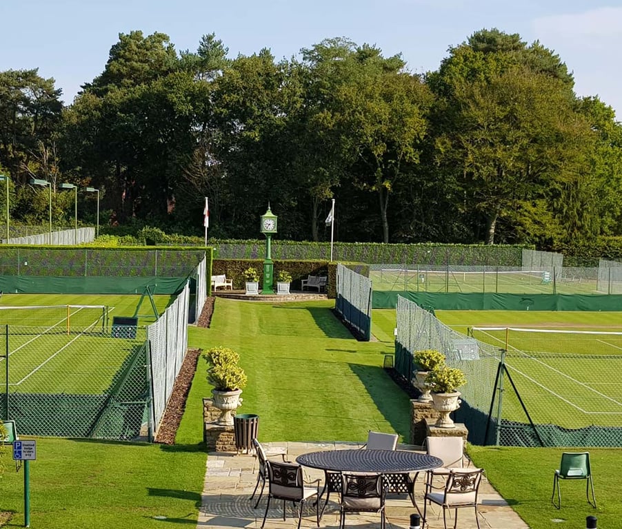 Scenic view of St George's Hill tennis courts with grass surfaces, surrounded by trees, and outdoor patio furniture in the foreground.