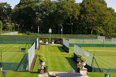 Scenic view of St George's Hill tennis courts with grass surfaces, surrounded by trees, and outdoor patio furniture in the foreground.