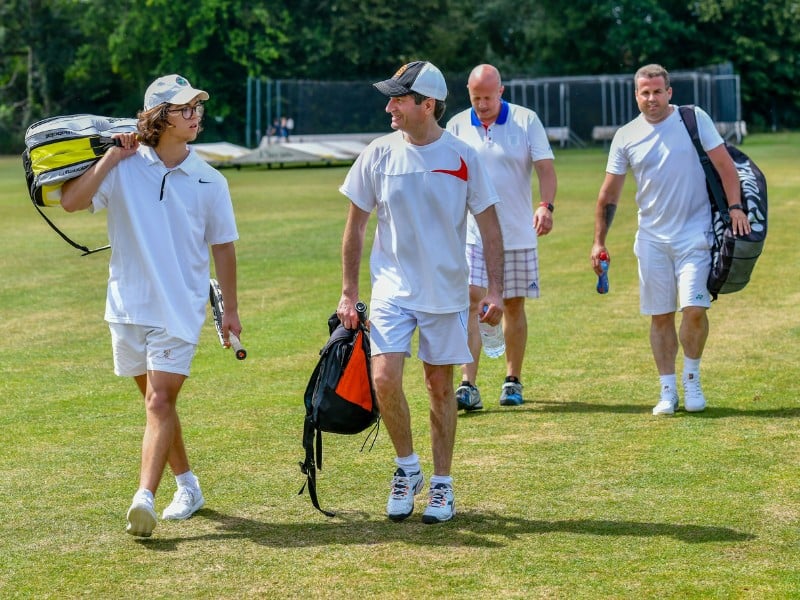Four men in sports attire walking on a cricket field, each carrying a cricket bag, with cricket nets visible in the background.