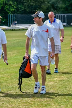 Four men in sports attire walking on a cricket field, each carrying a cricket bag, with cricket nets visible in the background.