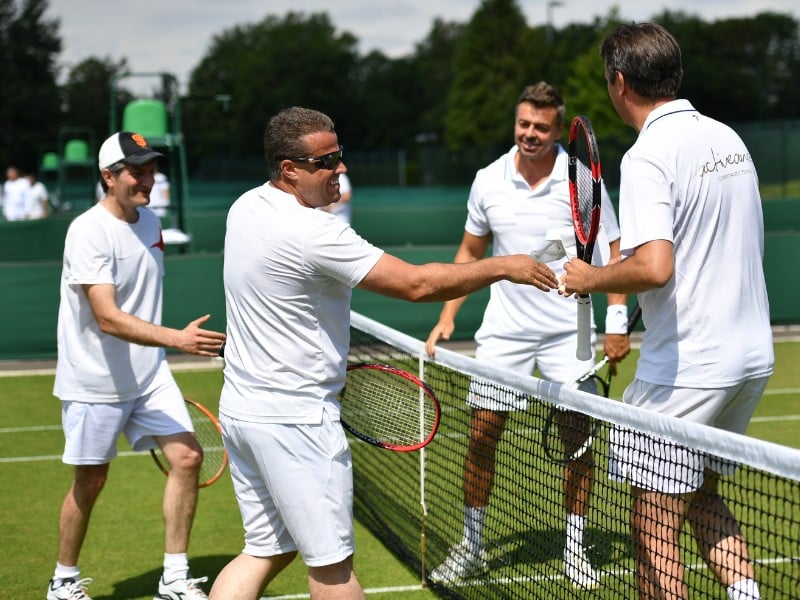 Four men in white tennis outfits shaking hands over the net on a grass court at St Georges Hill, showcasing sportsmanship.