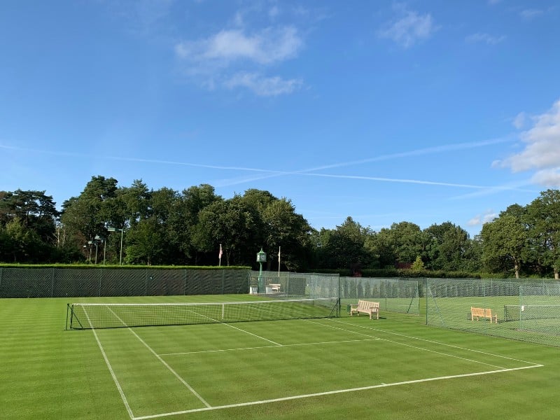 A grass tennis court at St Georges Hill Gallery, bordered by trees under a blue sky.