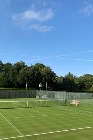 A grass tennis court at St Georges Hill Gallery, bordered by trees under a blue sky.