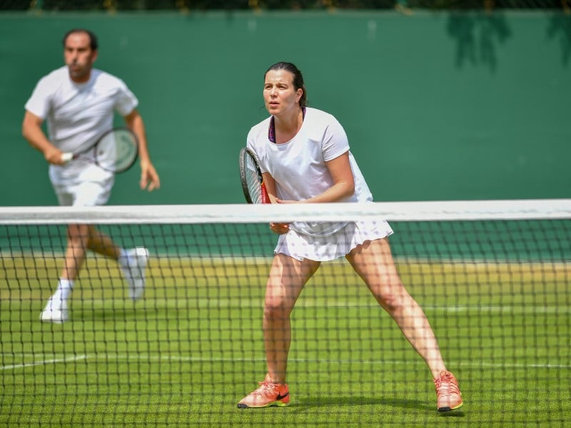 Mixed doubles tennis players at St Georges Hill; woman at the net in white attire with male partner in the background on a grass court.