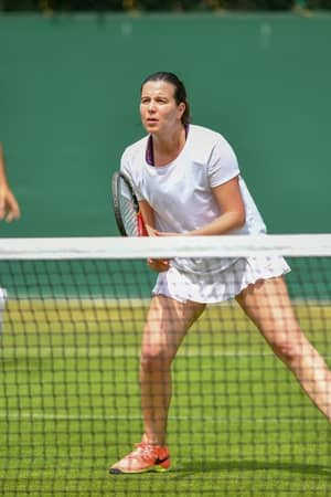 Mixed doubles tennis players at St Georges Hill; woman at the net in white attire with male partner in the background on a grass court.