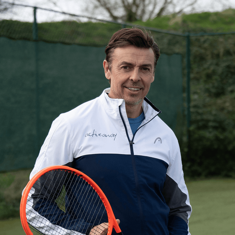 Steven Davies holding an orange tennis racket on a tennis court, wearing an Active Away jacket.