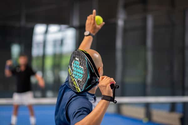 Male padel player serving on an indoor court, holding a padel racket and yellow ball, with another player in the background.