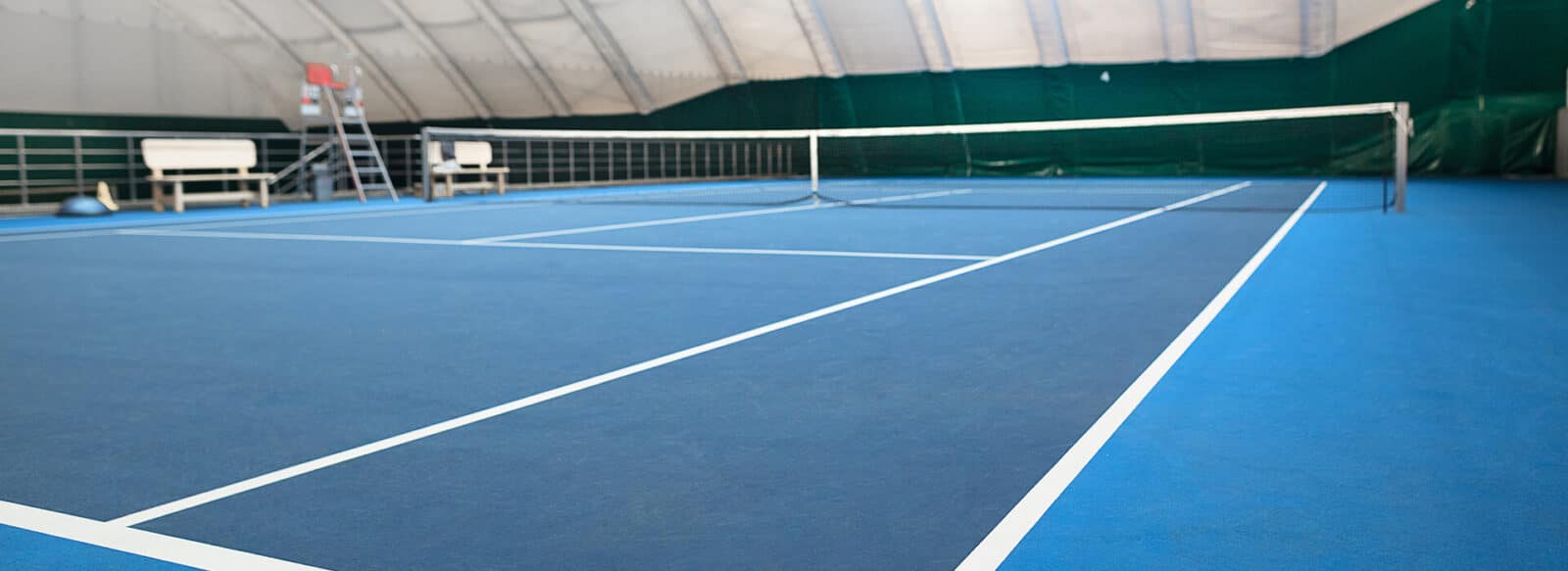 Indoor tennis court with blue surface and white lines, enclosed within a dome structure in the UK, used for tennis clinics in 2021.