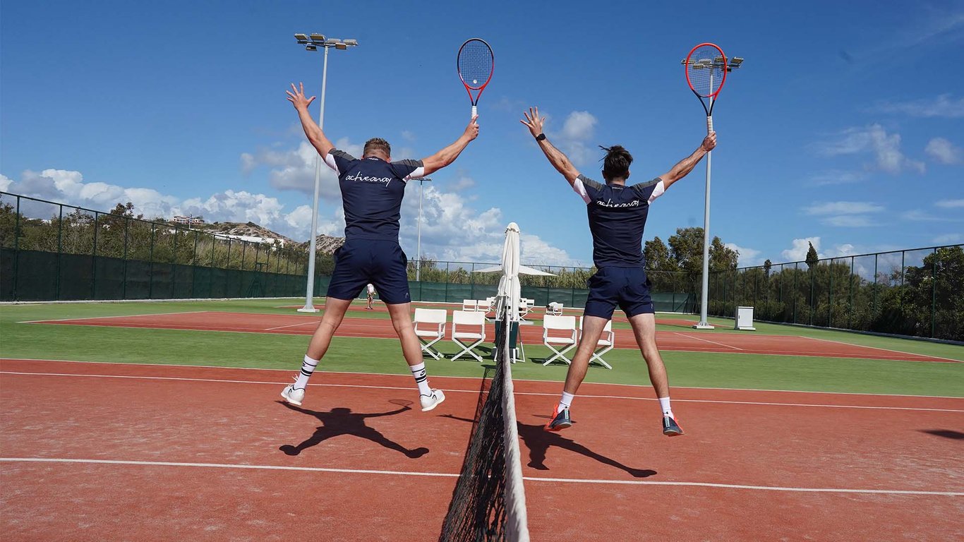Two male tennis players in navy sportswear jumping over the net on a clay court, holding rackets with a scenic background.