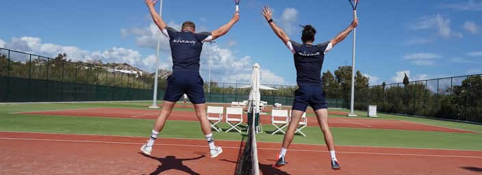 Two male tennis players in navy sportswear jumping over the net on a clay court, holding rackets with a scenic background.