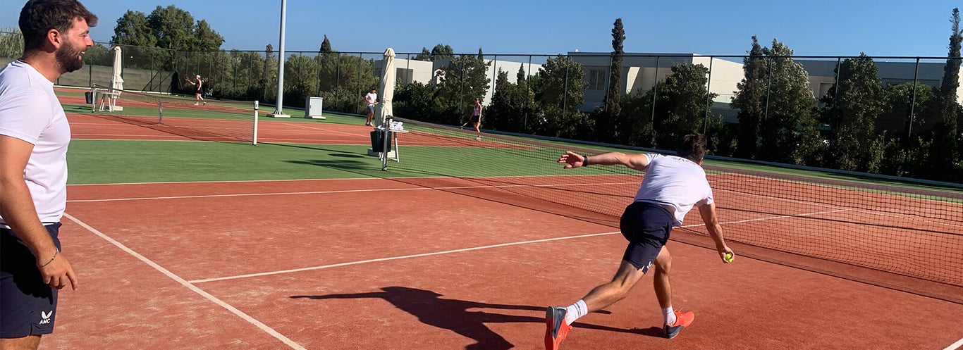 Two men warming up on a clay tennis court; one prepares to serve while the other watches.