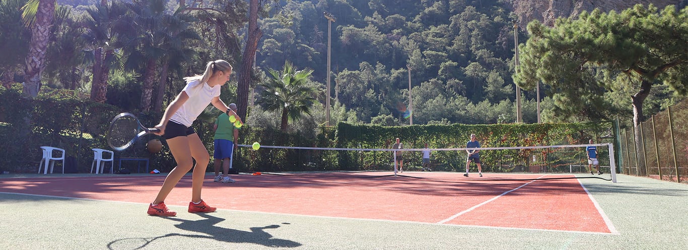 Young woman playing tennis on an outdoor court with lush green mountains in the background, part of the Autumn 2023 tennis holiday events.