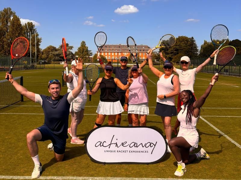 Active Away tennis team on grass courts at The Lensbury, posing with rackets raised, with the historic building in the background.