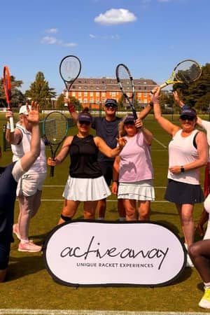 Active Away tennis team on grass courts at The Lensbury, posing with rackets raised, with the historic building in the background.